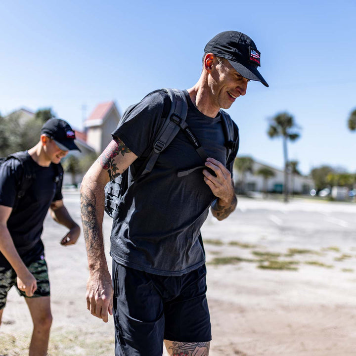 Two individuals, sporting GORUCK's Training Sternum Strap and black caps paired with athletic gear featuring MOLLE webbing, enjoy a sunny day outdoors.