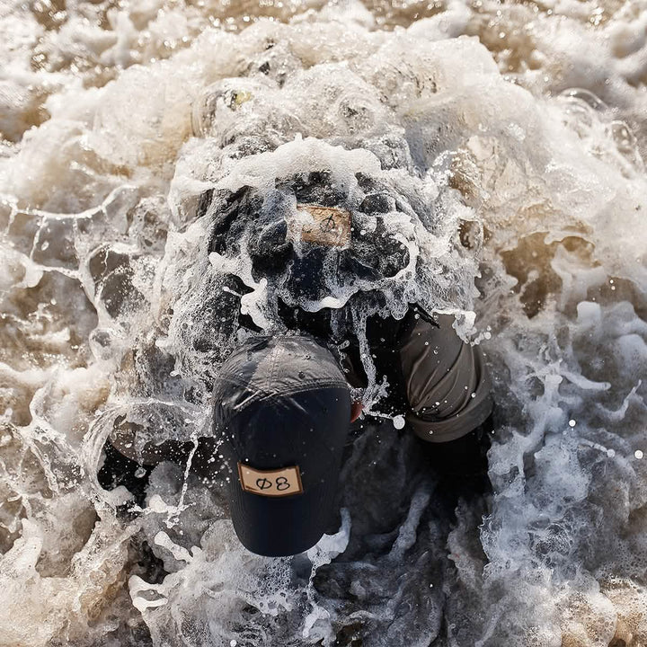 A person wearing the GORUCK Performance TAC Hat - TOUGHDRY is submerged in turbulent, foamy water. The hat, made from sweat-wicking ToughDry® fabric and featuring a distinct symbol, withstands the dynamic splashes that obscure parts of the individual.