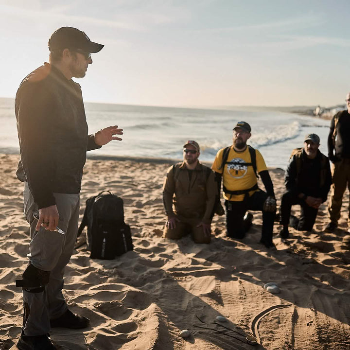 A man wearing a GORUCK Performance TAC Hat - TOUGHDRY and a dark jacket addresses a group kneeling on the sandy beach. The sunset elongates shadows over scattered backpacks and balls, as his sweat-wicking gear ensures comfort in the dimming light.