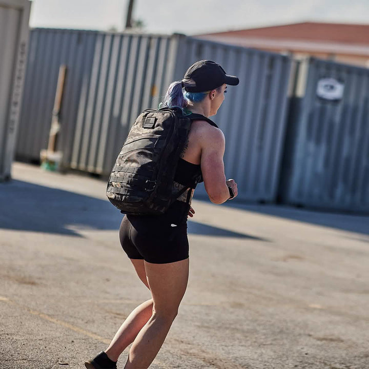 A person wearing a GORUCK Performance TAC Hat - TOUGHDRY in black, along with a tank top and shorts, is walking outdoors carrying a camouflage backpack. They're in an industrial area with large shipping containers in the background, equipped for any situation with gear crafted from durable ToughDry fabric.