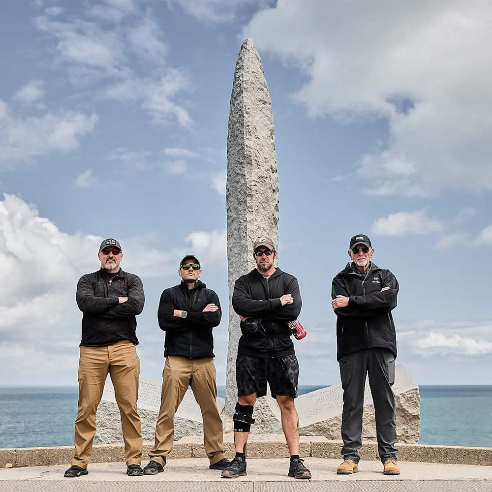 Four men stand with arms crossed in front of a tall, pointed monument by the sea. They are dressed in outdoor attire, including GORUCK's Performance TAC Hats made with TOUGHDRY material and sunglasses, under a partly cloudy sky. One man wears a knee brace and holds a water bottle.