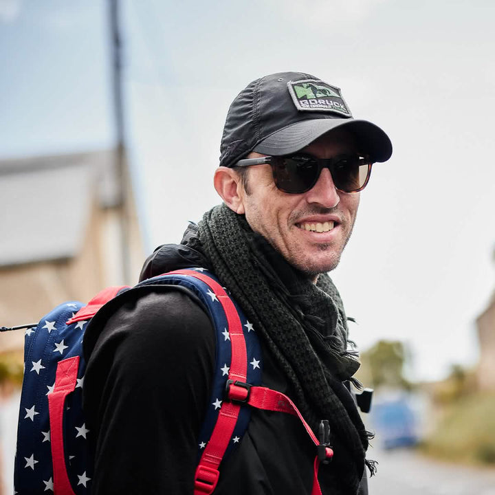 A person wearing the GORUCK Performance TAC Hat made with TOUGHDRY fabric smiles while standing outdoors, accessorized with sunglasses and a scarf. They carry a star-patterned backpack against the backdrop of a building under a clear sky, perfectly showcasing the hat's durable ToughDry® material and its sweat-wicking features.