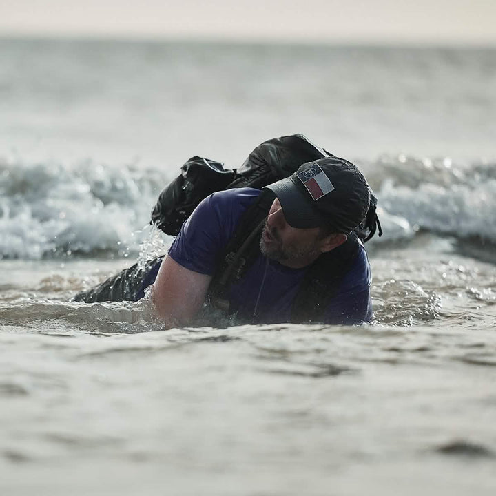 A person wearing a GORUCK Performance TAC Hat - TOUGHDRY and a backpack crawls through shallow ocean water as waves crash against them. The overcast sky enhances the scene's feeling of determination and challenge, while the TOUGHDRY fabric ensures they stay comfortable amidst nature's elements.