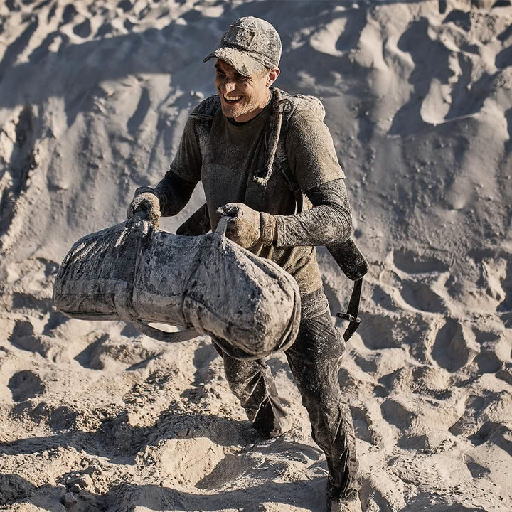 A person, wearing a GORUCK Performance TAC Hat - TOUGHDRY made from sweat-wicking material, climbs a sandy hill carrying a heavy sandbag. Clad in dark clothing and dirt-streaked, the scene evokes a grueling outdoor workout or demanding military training session.