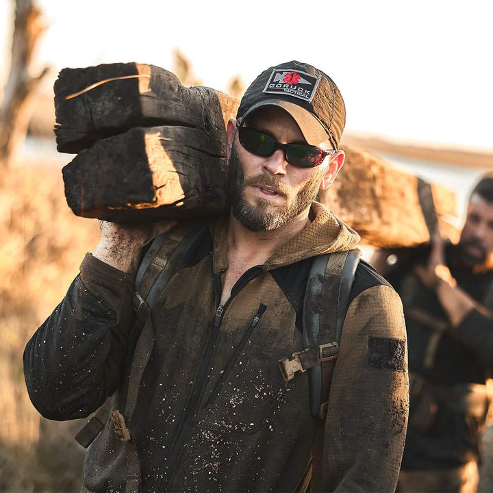 A man wearing sunglasses and a Performance TAC Hat from GORUCK, built with TOUGHDRY fabric, carries large wooden beams on his shoulder while clad in a dirt-covered jacket. The scene takes place outdoors, accompanied by another person carrying similar beams, emphasizing the toughness and resilience of TOUGHDRY fabric during manual labor or outdoor work.