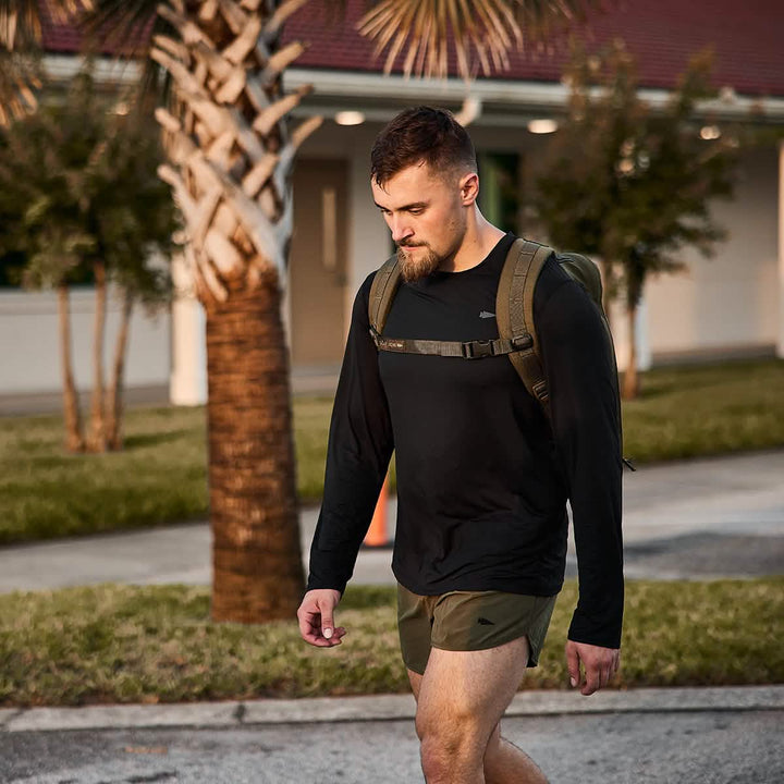 A bearded man, wearing a GORUCK Men's Long Sleeve Performance Tee in black and green shorts, strolls outside with a backpack. Palm trees and a building provide the backdrop under the sunlight, reminiscent of walks in the Italian Alps.