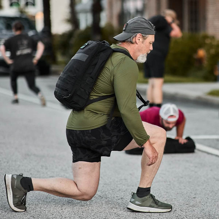 A man wearing the Men's Long Sleeve Performance Tee - ToughMesh by GORUCK and black shorts, with a backward cap, is participating in outdoor physical activity. He is lifting or moving an item with both hands, with a palm tree and building in the background.