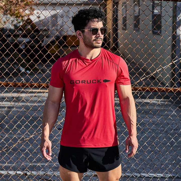 A man in a Men’s Performance Tee - ToughMesh by GORUCK, featuring a bold red color, sunglasses, and black shorts stands in front of a chain-link fence. The sun casts shadows on the ground behind him, highlighting the shirt's quick-drying performance and featherweight design.