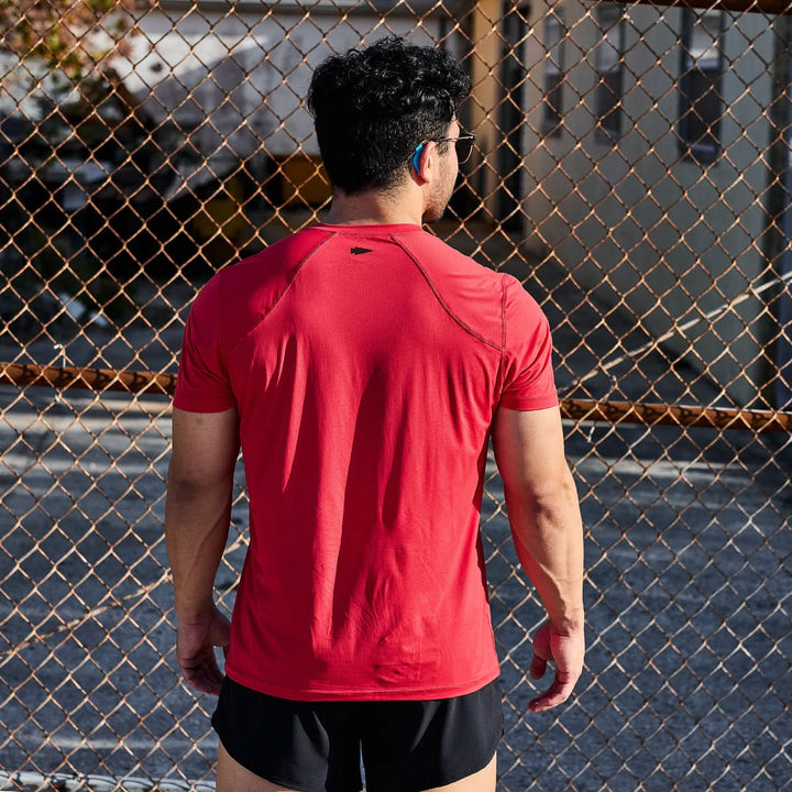 A person wearing a Men’s Performance Tee - ToughMesh from GORUCK and black shorts stands with their back to the camera, facing a chain-link fence under sunlight.