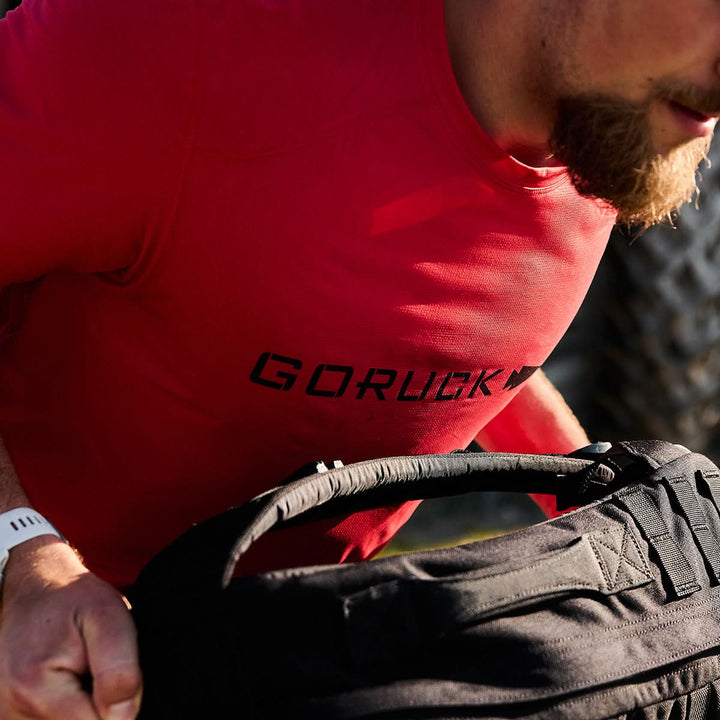 A person with a beard is wearing a red GORUCK Men’s Performance Tee - ToughMesh and carrying a black backpack outdoors. The sunlight casts shadows on the quick-drying fabric of the tee.