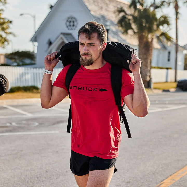 A man wearing a GORUCK Men’s Performance Tee - ToughMesh and black shorts is walking on a street, carrying a heavy backpack on his shoulders. A white church and palm trees are in the background under a clear sky.