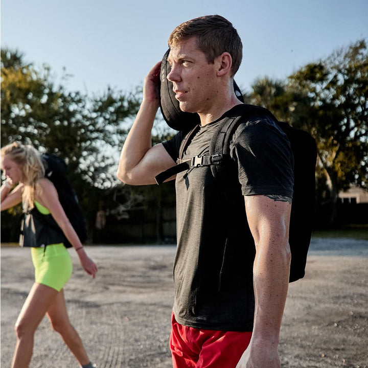 A man in a Men’s Performance Tee - ToughMesh by GORUCK and red shorts carries a backpack over his shoulder while standing outdoors. A woman in green shorts and a black top walks beside him, with trees and a clear sky, reminiscent of the serene Italian Alps, in the background.