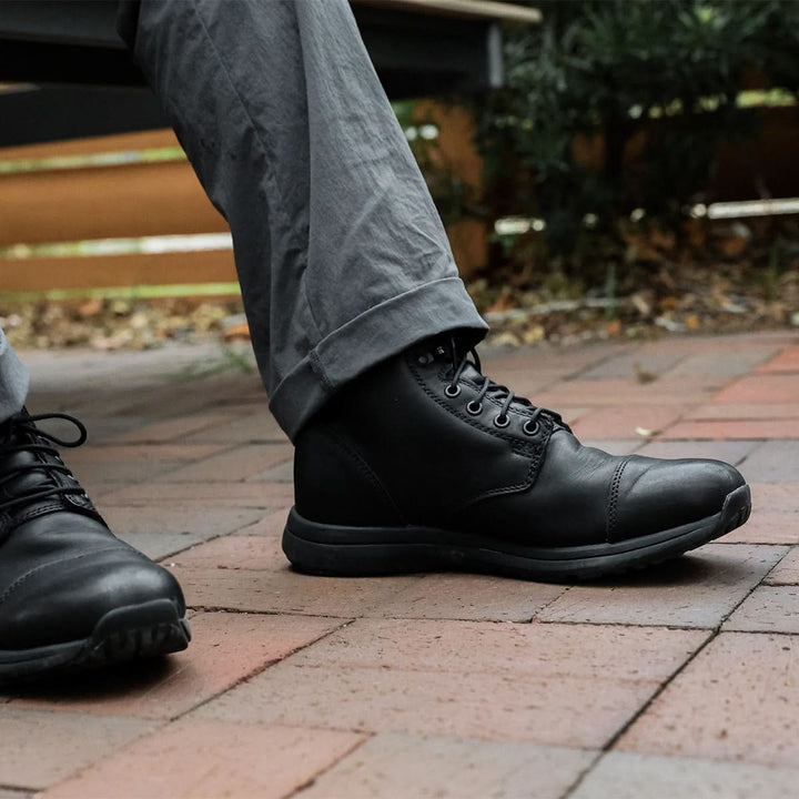 Close-up of a person wearing GORUCK's MACV-1 Traveler boots, featuring an all-terrain outsole, paired with gray pants while standing on a red brick surface. The background includes a wooden bench and some greenery.