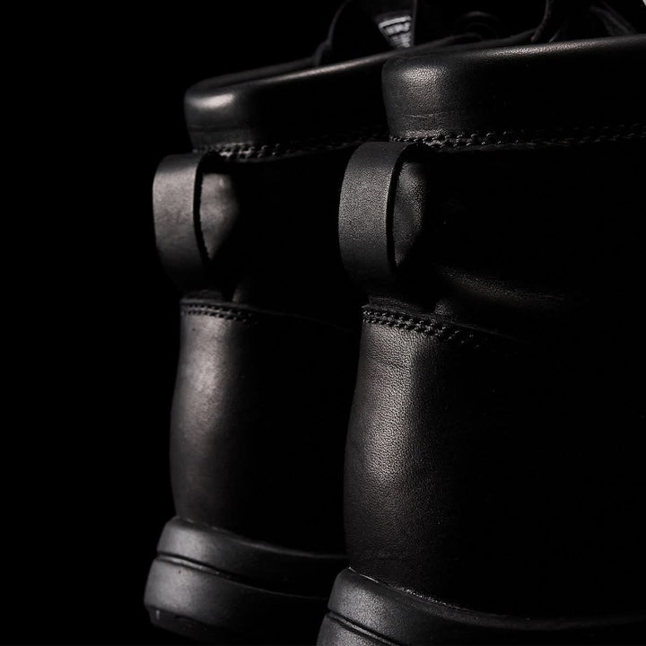 Close-up of the back of two black leather boots against a dark background. The MACV-1 Traveler boots by GORUCK feature padded collars and a sleek design with an all-terrain outsole, emphasizing their rugged and sturdy appearance.