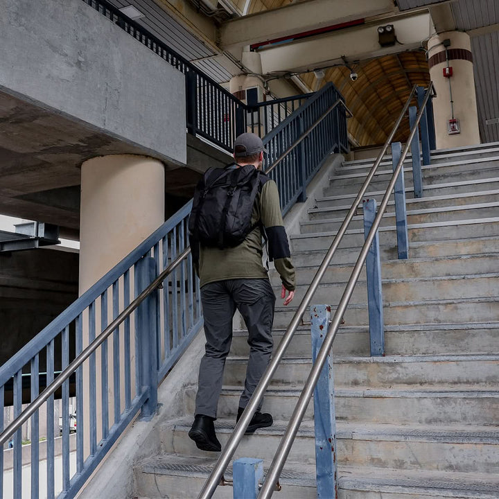 A person wearing a backpack, green jacket, gray pants, and a cap strides up a flight of concrete stairs in their MACV-1 Traveler footwear by GORUCK. The metal railings and visible beams under the sloped roof complement the tough, reliable grip of their All-Terrain Outsole.