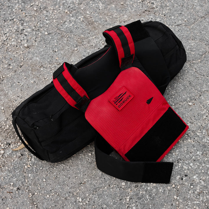 A black sports bag and a GORUCK Training Weight Vest 2.0 in red, ideal for weight vest exercises, rest on a gravel surface.