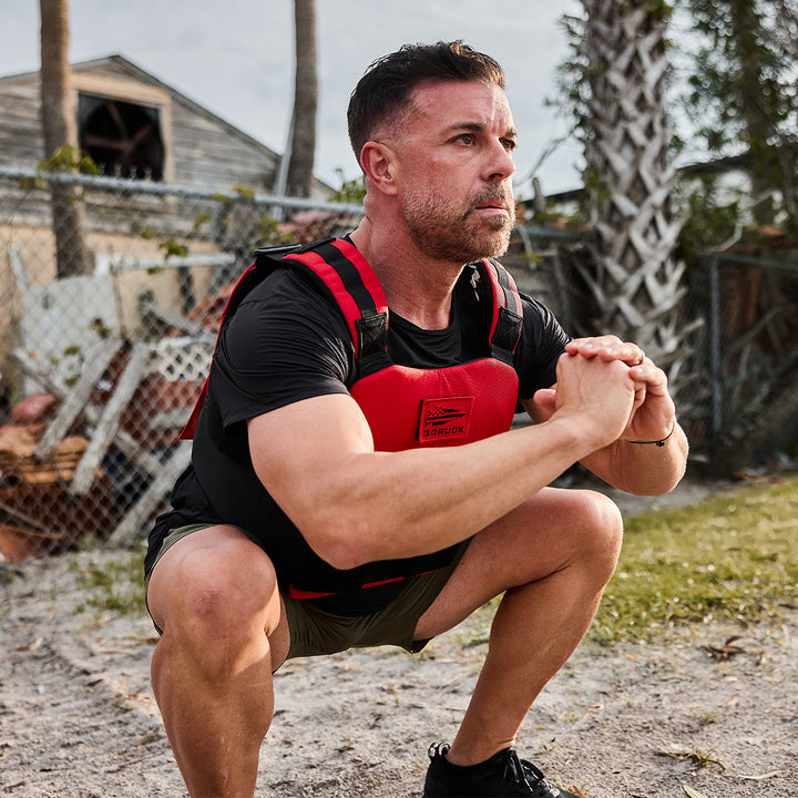 Wearing a GORUCK Training Weight Vest 2.0, a man performs squats outdoors near a chain-link fence, surrounded by swaying palm trees.