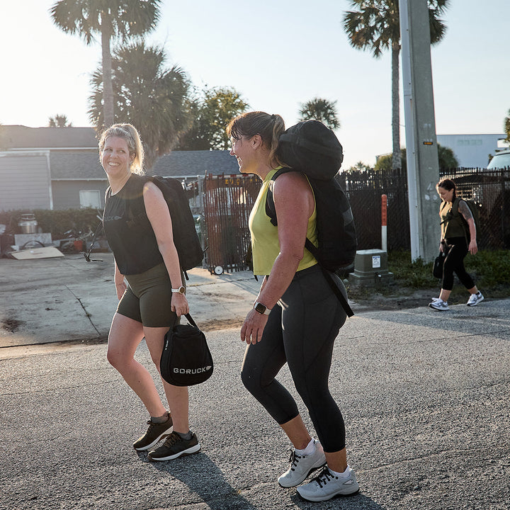 Two women are walking on a sunny street, smiling while wearing athletic clothing and backpacks. One carries a GORUCK black bag made from durable CORDURA® Ballistic Nylon. In the background, another person walks near a fence, with palm trees swaying gently above them as they wear Women's Ballistic Trainers in Lunar Rock + Charcoal by GORUCK, featuring the Silver Reflective Spearhead.