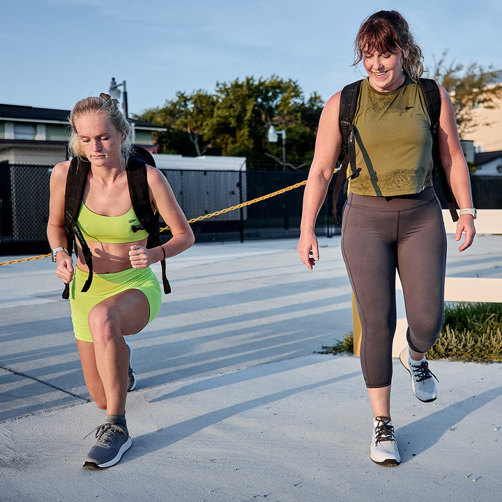 Two women exercising outdoors with weighted vests capture the essence of functional fitness. One, wearing a vibrant yellow workout set and GORUCK's Women's Ballistic Trainers in Lunar Rock + Charcoal with Silver Reflective Spearhead, lunges gracefully beside her companion. She is walking in a green top and gray leggings along a paved path with trees gently swaying behind them.