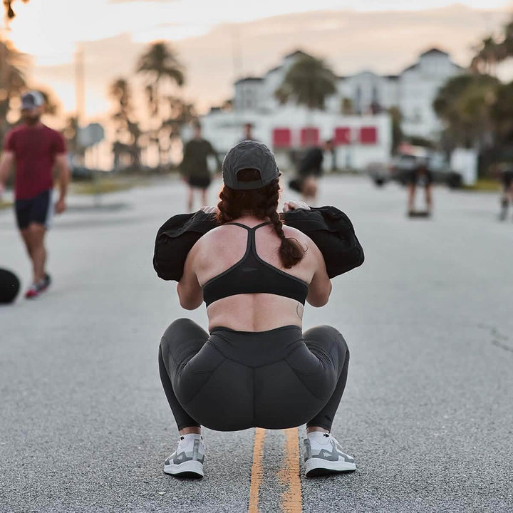 A woman squats on the street, exuding strength in GORUCK Women's Training Leggings Pocket - ToughFlex, which are crafted from durable, squat-proof fabric. Surrounded by an urban backdrop, she showcases determination and style in her workout gear.