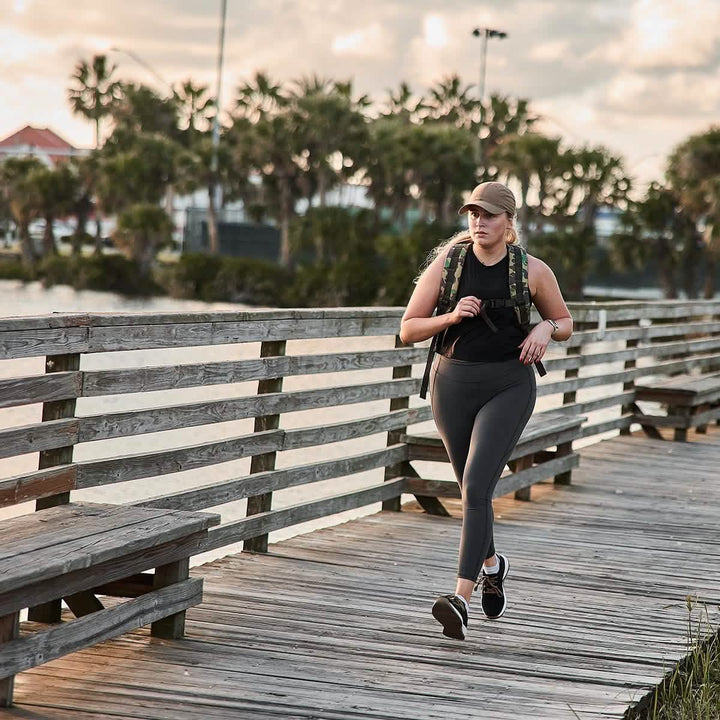 A woman walks along a wooden boardwalk by the water, dressed in athletic wear and a cap. Her GORUCK Women's Training Leggings Pocket, made from ToughFlex Fabric, ensure they are squat-proof. Tall palm trees sway gently in the background.