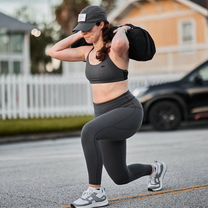 Wearing GORUCK Women's Training Leggings Pocket - ToughFlex, a woman lunges with a sandbag outdoors. In her sleek workout gear and cap, she embodies strength and resilience as she powers through her routine.