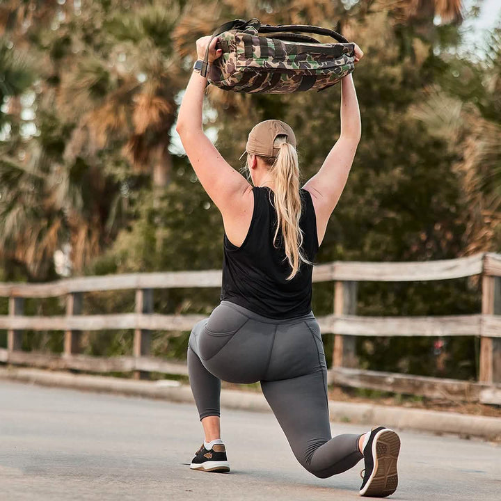 A person in athletic wear, donning GORUCK Women’s Training Leggings Pocket - ToughFlex, lunges on a path with a camo bag overhead. The squat-proof outfit complements the serene backdrop of trees and a wooden fence.