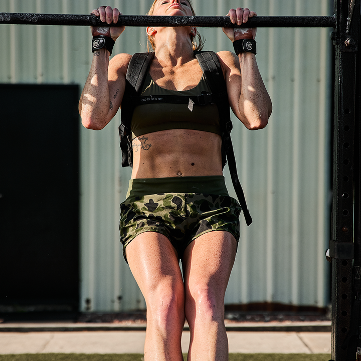 Outside the building, a person does a pull-up on a sturdy metal bar wearing GORUCK Women’s Training Shorts - ToughStretch and carrying a backpack.