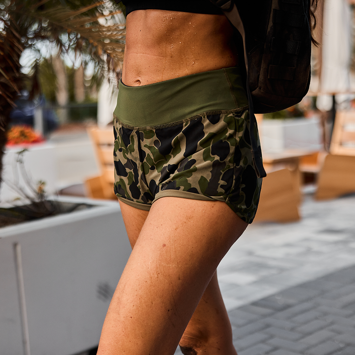 A person in camo shorts and a black top confidently strides outdoors on a sunny day, wearing GORUCK Women's Training Shorts - ToughStretch for ultimate comfort and flexibility.