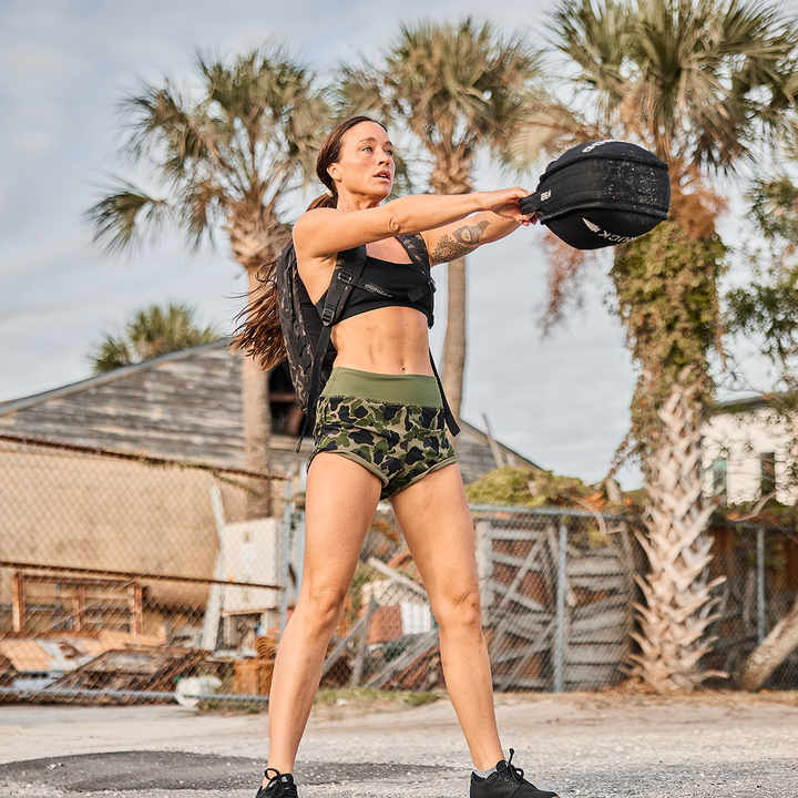 A woman exercises outdoors with a sandbag, confidently wearing GORUCK’s squat-proof Women’s Training Shorts - ToughStretch amidst palm trees and a rustic building, showcasing her strength and style.