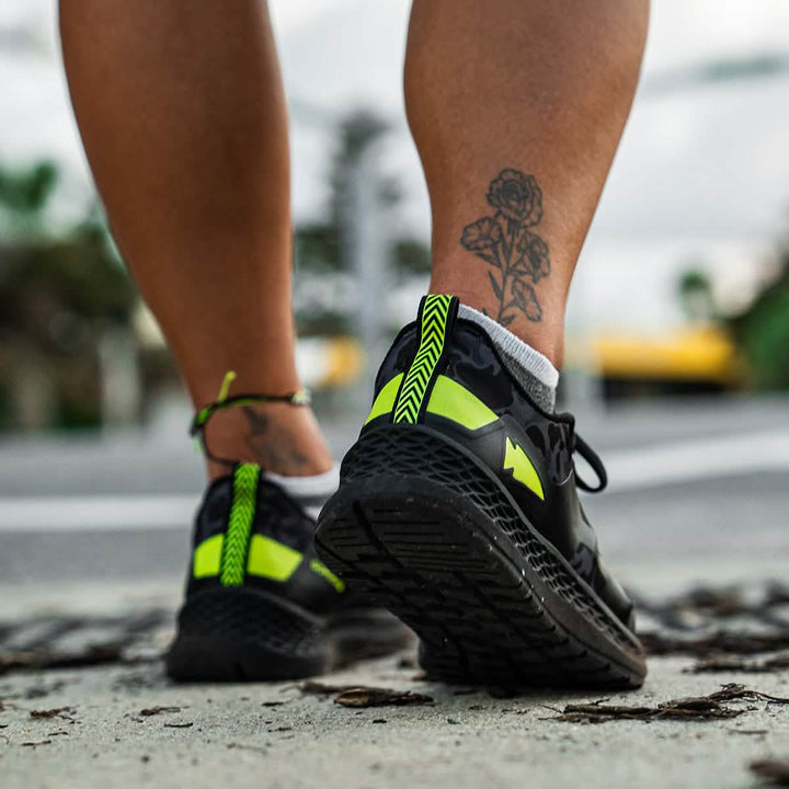 A person strides down the sidewalk in GORUCK's Rough Runner - Midnight Frogskin + Acid Lime sneakers, which feature a versatile road-to-trail outsole and gradient density EVA midsole for comfort. A rose tattoo on their calf captures attention just as much as their footwear.