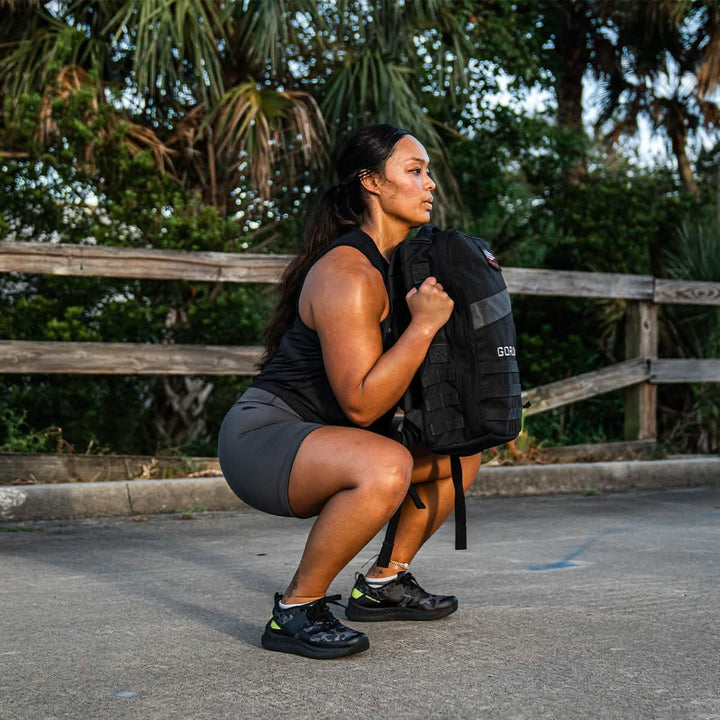 A woman in athletic wear squats outdoors with her black backpack, surrounded by palm trees. Her GORUCK Rough Runner shoes in Midnight Frogskin + Acid Lime feature a versatile road-to-trail outsole, perfect for any adventure she's planning to tackle.