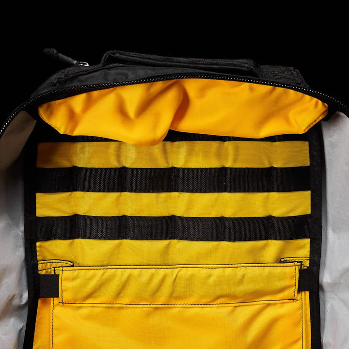 The GORUCK GR1 USA - X-PAC backpack, showcased against a black background, sports a yellow interior and black horizontal straps. Crafted from durable X-PAC fabric with AquaGuard zippers, it's waterproof and designed for adventurous journeys.