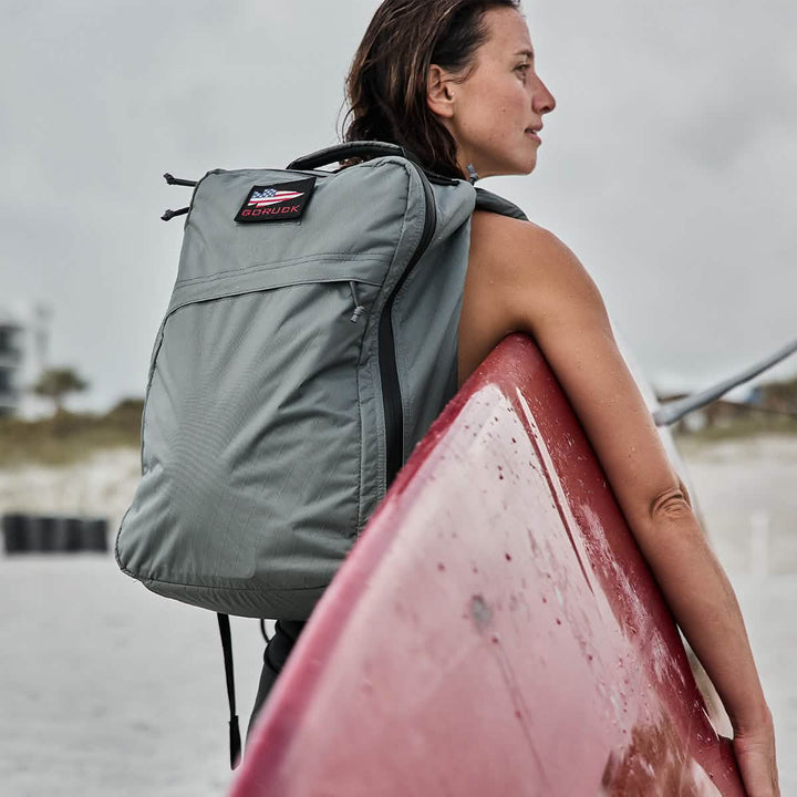 A person holds a red surfboard and wears a GORUCK GR1 USA - X-PAC backpack featuring AquaGuard zippers as they stand on the beach under a cloudy sky.