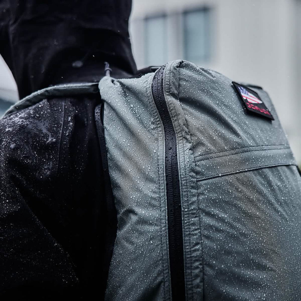 In an urban setting, a person strolls with a GR1 USA - X-PAC backpack by GORUCK and a rain jacket featuring AquaGuard zippers, all covered in raindrops.