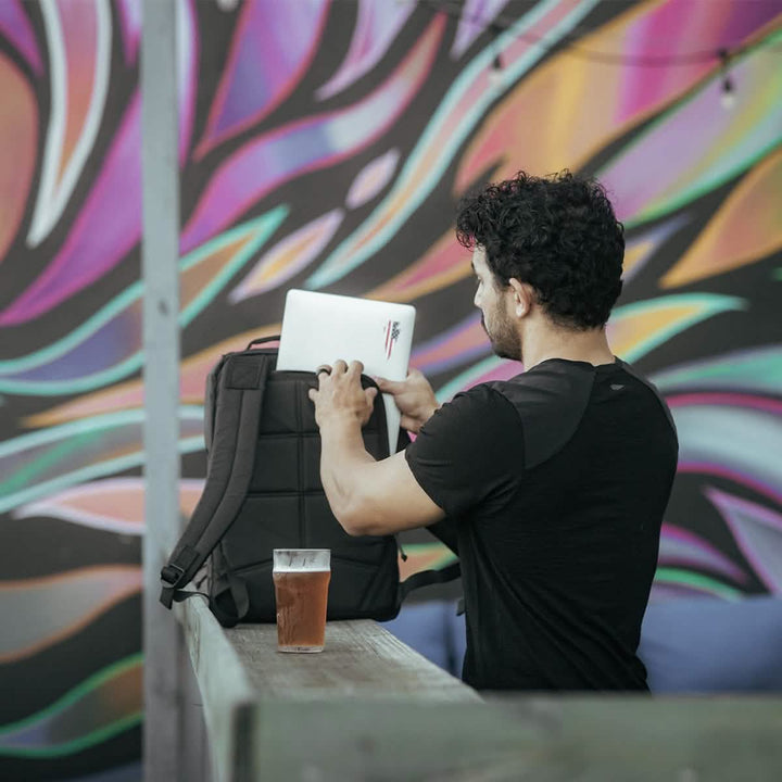 A man is placing a laptop into a GR1 USA - X-PAC by GORUCK, which features waterproof AquaGuard zippers, on a table with a glass of beer; a colorful mural paints the background.