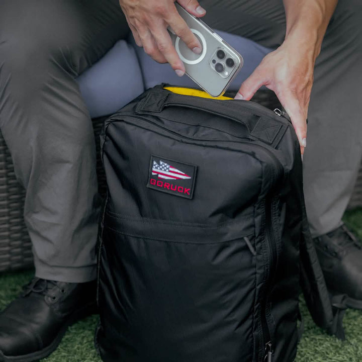 A person is placing a smartphone into the open pocket of a sleek black GR1 USA - X-PAC backpack by GORUCK while sitting on a gray cushion.