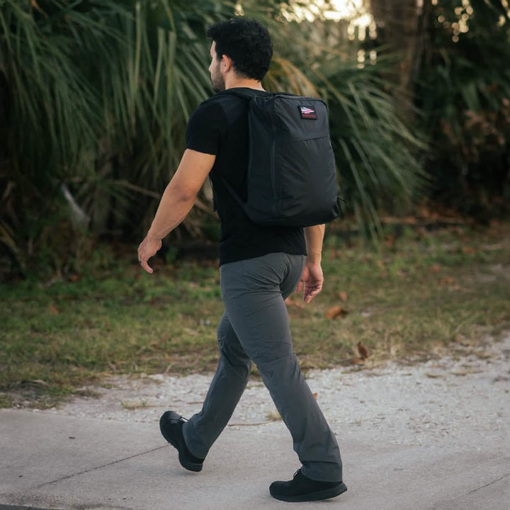 A man strolls along the sidewalk, sporting a GORUCK GR1 USA - X-PAC backpack with AquaGuard zippers, wearing a black shirt and gray pants, surrounded by lush greenery.