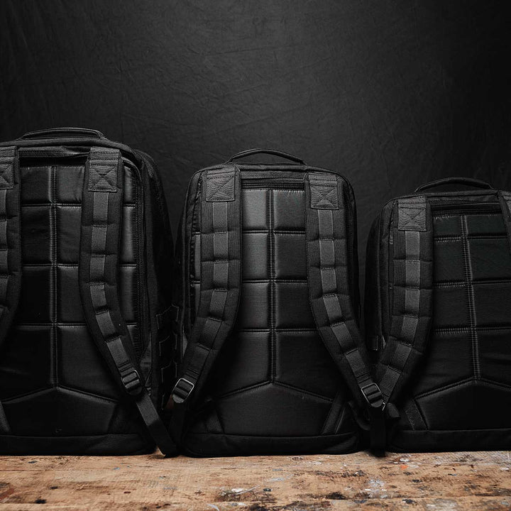 Three black GR2 - USA backpacks by GORUCK, featuring padded straps and crafted from durable 210D CORDURA®, are positioned on a wooden surface against a dark background. Each backpack is carry-on compliant, ensuring a perfect blend of style and convenience for your travel needs.