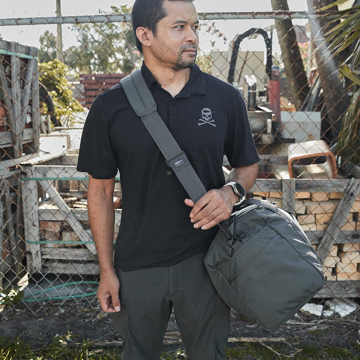 A person stands outdoors in front of a chain-link fence, wearing a black polo shirt with a skull logo and carrying a GORUCK Kit Bag (Includes Shoulder Strap). The background includes stacked wooden pallets and trees.