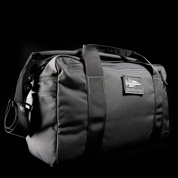 Set against a dark background, the Kit Bag by GORUCK is a sleek black duffel featuring sturdy handles, a shoulder strap, and a logo-adorned patch on the side. Perfect for adventurers, this bag combines functionality with style for any journey.