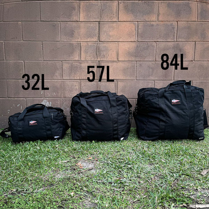 Three black Kit Bags by GORUCK, featuring different sizes, are artfully arranged outdoors against a brick wall. Labeled 32L, 57L, and 84L to indicate their capacities, these rugged bags rest on the grass with their included shoulder straps, ready for any adventure that calls for an aviator’s kit bag.