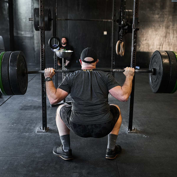 In a dimly lit gym filled with weightlifting equipment, a person performs a squat with a barbell, highlighting their Men's Ballistic Trainers - Black + Gum w/ Black Reflective Spearhead from GORUCK. Nearby, another individual sits in the background, either observing or taking a break.