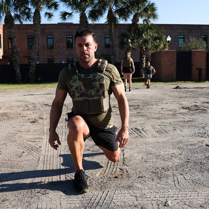 A man dressed in a camouflaged vest and black shorts, sporting GORUCK's Men's Ballistic Trainers in Black + Gum with a Black Reflective Spearhead, performs a lunge exercise outdoors on a dirt surface. In the background, two more individuals can be seen exercising amidst palm trees and a brick building under the clear sky.