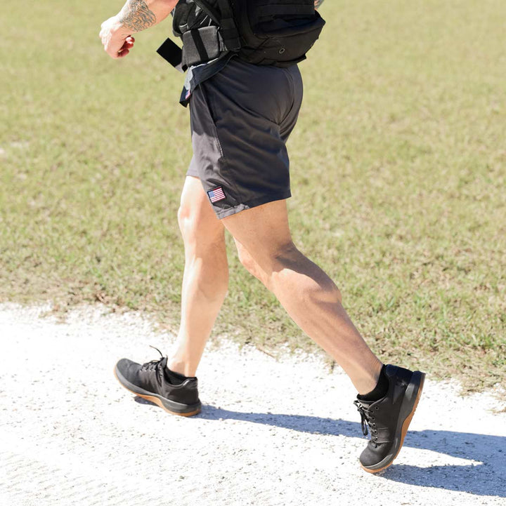 A person with a tattoo on their arm strolls along a gravel path, sporting a black backpack and gray shorts adorned with a small American flag. They wear GORUCK Men's Ballistic Trainers - Black + Gum w/ Black Reflective Spearhead, crafted with durable CORDURA® Ballistic Nylon, suggesting functional fitness against the backdrop of a grassy field.