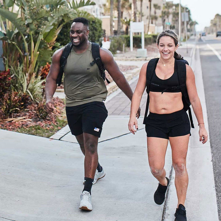 Two people in athletic gear, sporting GORUCK Ballistic Trainers in Lunar Rock, stroll along a sunny sidewalk with smiles and backpacks.