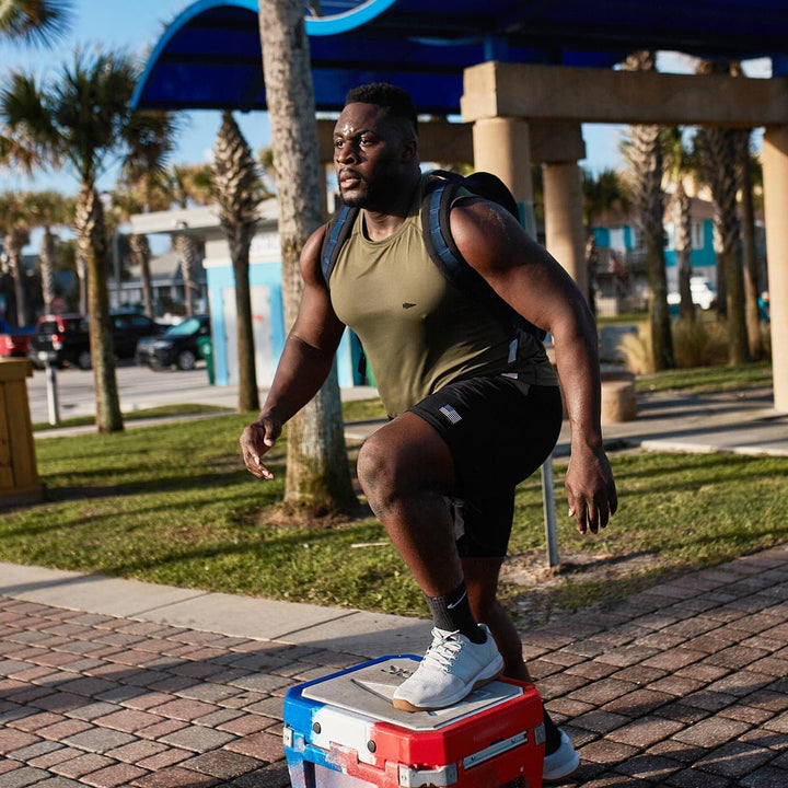 A man exercises outdoors in GORUCK's Ballistic Trainers - Lunar Rock, stepping on a cooler while carrying a backpack crafted from CORDURA® Ballistic Nylon, surrounded by swaying palm trees.