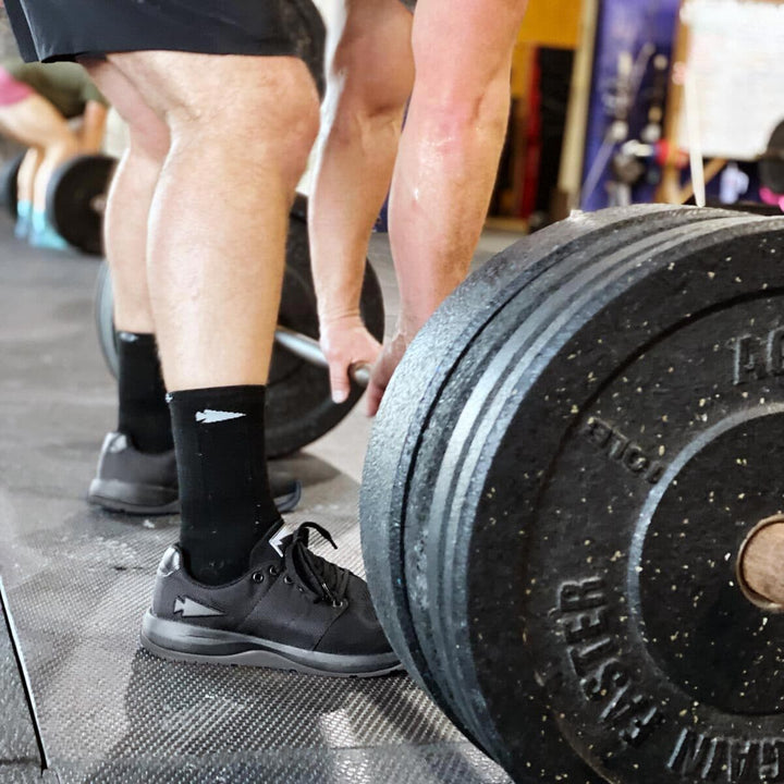 In the gym, a person wearing GORUCK Ballistic Trainers in Blackout with Black Reflective Spearhead lifts a heavy barbell, their shoes and socks providing a solid foundation. Others exercise in the background, inspired by the focus and strength on display.