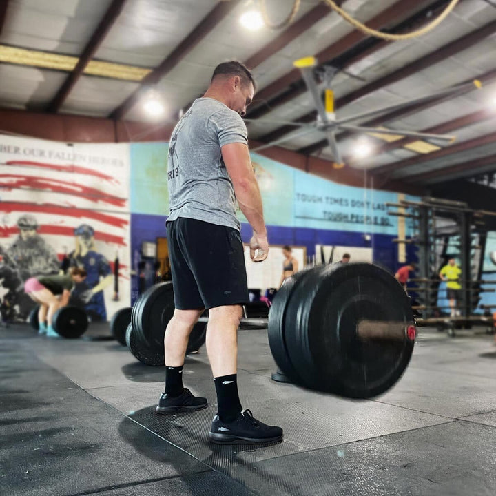 A man, focused and determined, lifts heavy weights in the gym, his Ballistic Trainers - Blackout w/ Black Reflective Spearhead by GORUCK gripping the floor beneath an American flag mural. The shoes' CORDURA® Ballistic Nylon laces promise durability as he strives for peak performance with their 3X Support™.