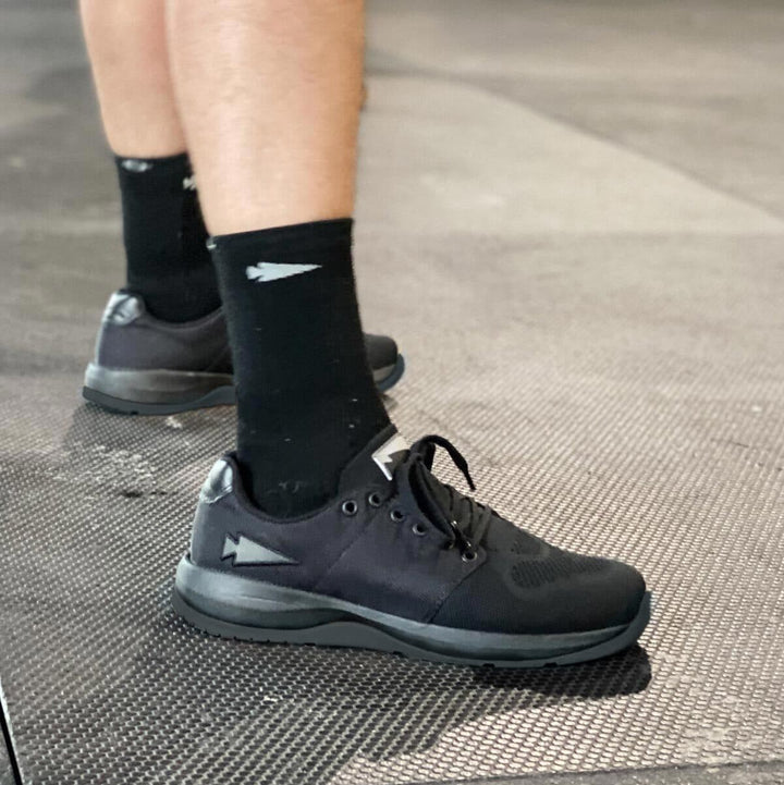 An individual wearing the Ballistic Trainers - Blackout with Black Reflective Spearhead from GORUCK, paired with socks featuring a geometric pattern, stands on a textured floor, highlighting this functional fitness footwear designed for optimal performance.